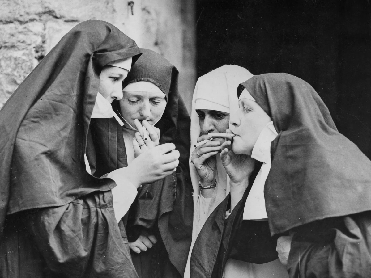 Four nuns in dark hoods and habits gathered closely, with all holding a cigarette.